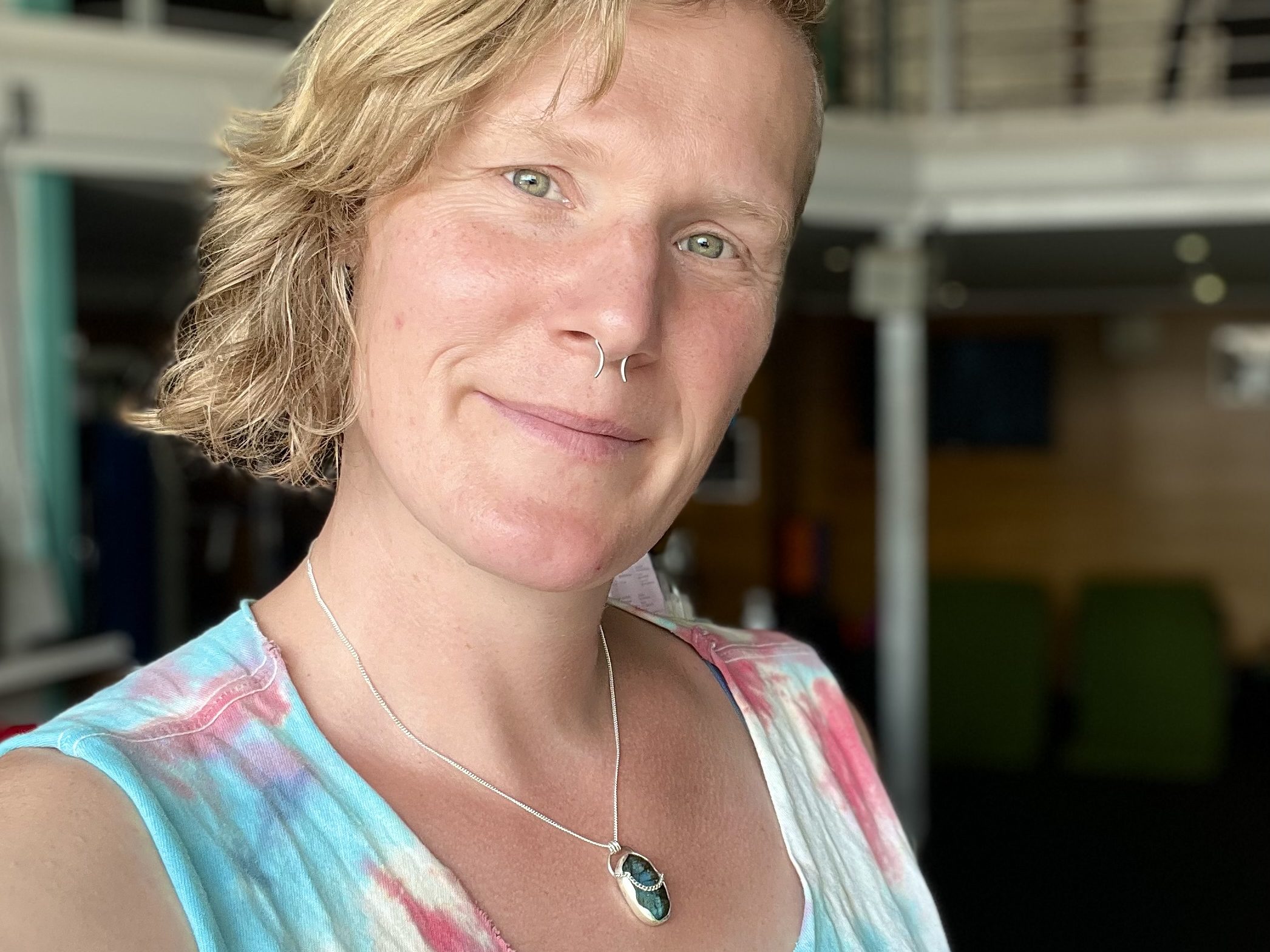 Huld, a white femme, smiles at the viewer. She has medium long blonde wavy hair and is wearing a pendant with a blue-hued irregular stone and a blue and pink sleeveless top.